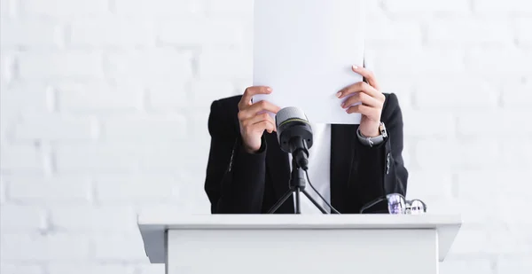 Young lecturer, suffering from fear of public speaking, standing on podium tribune and hiding face behind paper sheet — Stock Photo