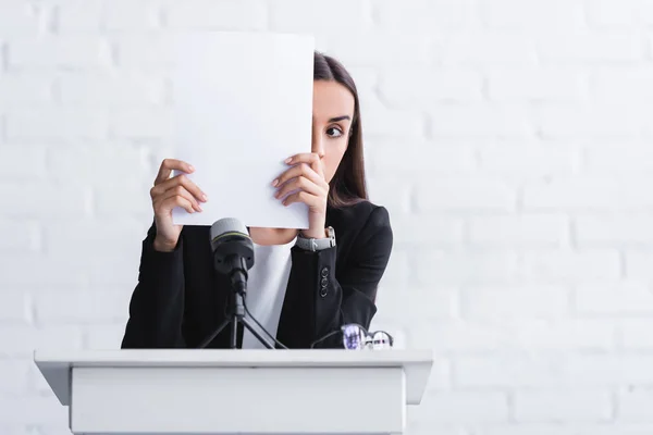 Young lecturer suffering from fear of public speaking hiding face with paper sheet while standing on podium tribune — Stock Photo