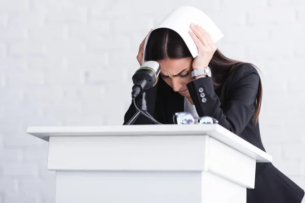 Young lecturer, suffering from fear of public speaking, standing on podium tribune and covering head with paper — Stock Photo