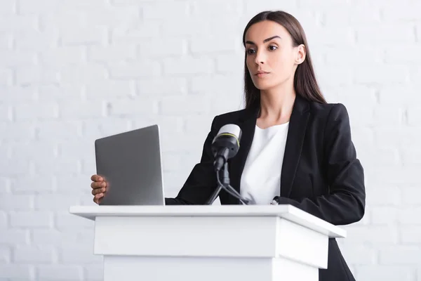 Attractive, confident lecturer standing on podium tribune near microphone and laptop — Stock Photo