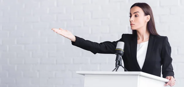 Panoramic shot of beautiful, serious lecturer pointing with hand while standing on podium tribune — Stock Photo