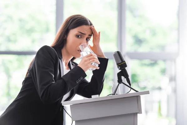 Pretty young lecturer, suffering from glossophobia, drinking water and holding hand near face while standing on podium tribune — Stock Photo