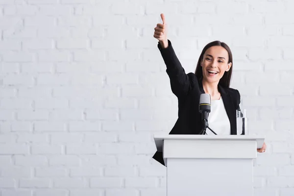 Happy, attractive lecturer showing thumb up while standing on podium tribune — Stock Photo