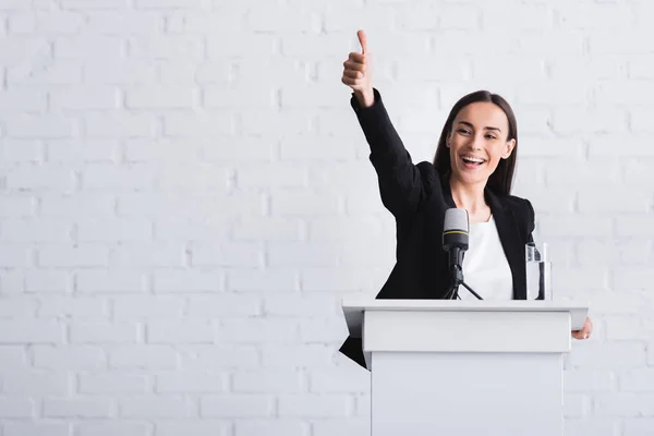 Feliz, atractivo conferenciante mostrando el pulgar hacia arriba mientras está de pie en el podio tribuna - foto de stock