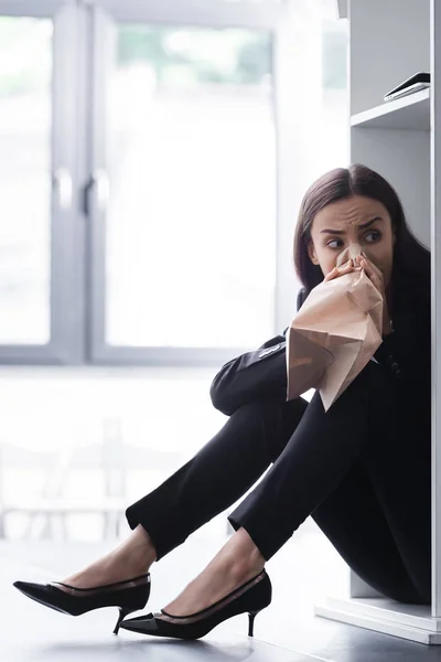 Scared lecturer sitting on floor in conference hall while suffering from panic attack and breathing into paper bag — Stock Photo