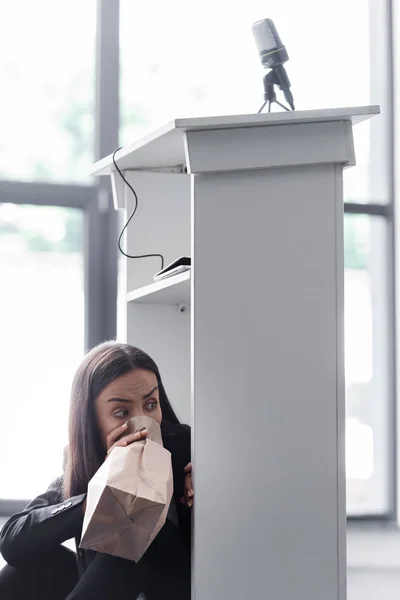 Испуганный лектор дышит в бумажный пакет, сидя на полу в конференц-зале и страдая от панической атаки — стоковое фото