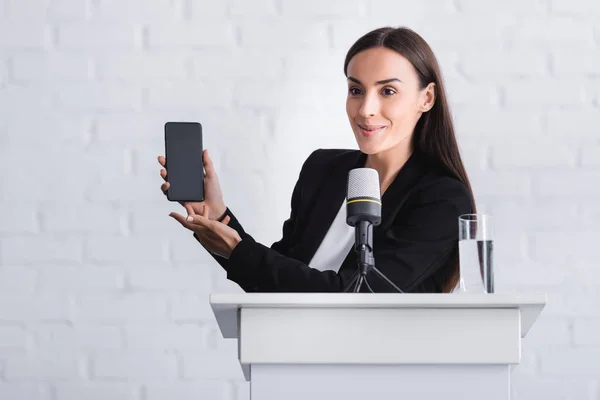 Palestrante alegre em pé no tribuno pódio e apresentando smartphone com tela em branco — Fotografia de Stock