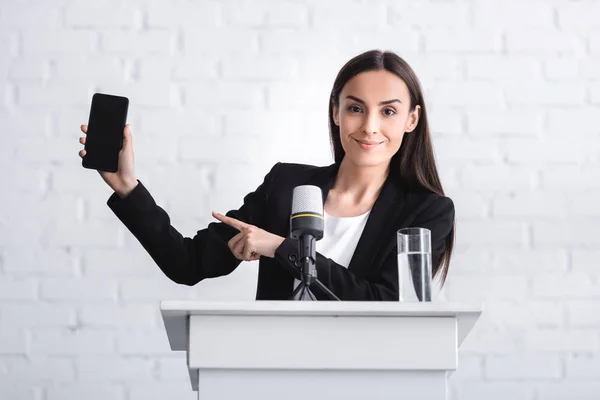 Sonriente conferenciante de pie en tribuna podio y apuntando con el dedo al teléfono inteligente con pantalla en blanco - foto de stock