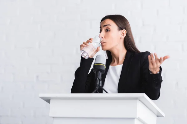 Jolie, jeune conférencier boire de l'eau et gestuelle tout en se tenant sur le podium tribune — Photo de stock