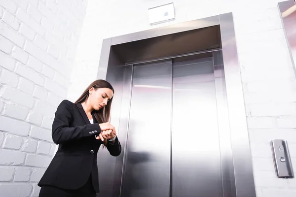 Joven mujer de negocios en el tiempo de control de desgaste formal de guardia mientras espera el ascensor - foto de stock