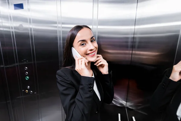 Cheerful businesswoman talking on smartphone in elevator and smiling at camera — Stock Photo