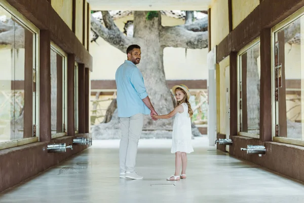 Happy father holding hands with cheerful and cute daughter in straw hat and white dress while standing in zoo — Stock Photo