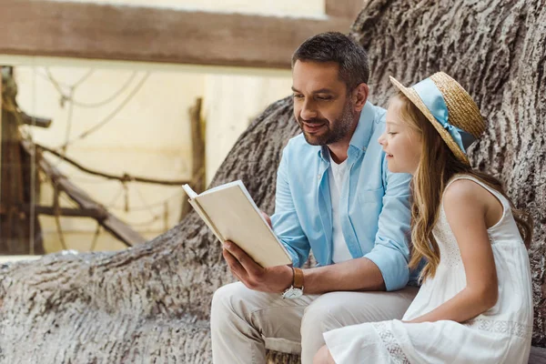 Веселий батько читає книгу біля милої дочки в солом'яному капелюсі біля дерева — стокове фото