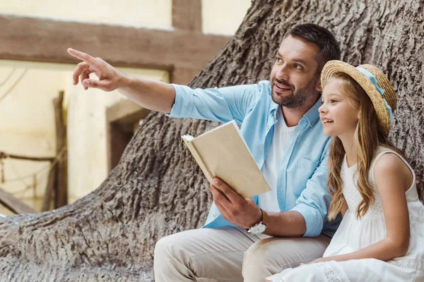 Cheerful father holding book and pointing with finger near cute daughter in straw hat — Stock Photo