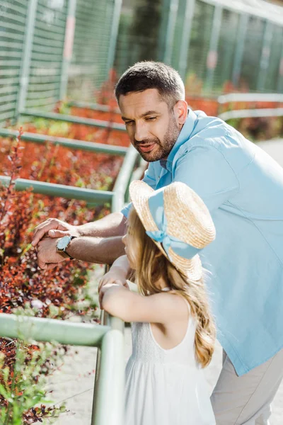 Hombre guapo mirando a la hija en sombrero de paja y vestido de pie en el zoológico - foto de stock