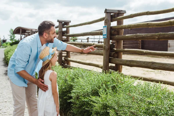 Hombre feliz señalando con el dedo cerca de la hija en sombrero de paja y vestido mientras está de pie en el zoológico - foto de stock
