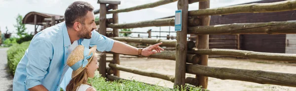 Colpo panoramico di uomo felice che punta con dito vicino figlia in cappello di paglia mentre in piedi nello zoo — Foto stock