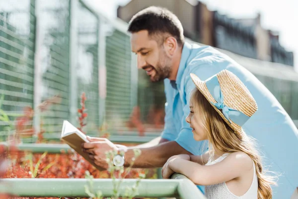 Enfoque selectivo de niño en sombrero de paja de pie cerca de padre guapo libro de lectura - foto de stock