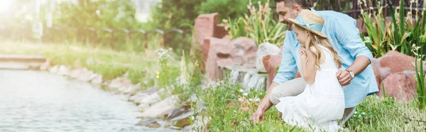 Tiro panorámico de lindo niño en vestido y sombrero de paja sentado cerca de feliz padre y el lago - foto de stock