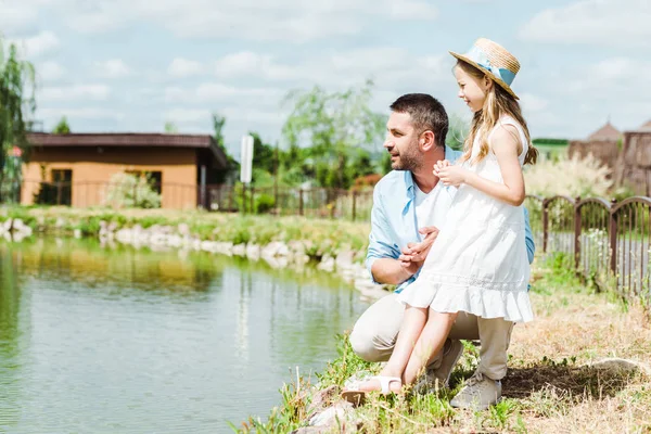 Mignon enfant en robe et chapeau de paille debout près heureux père et lac — Photo de stock