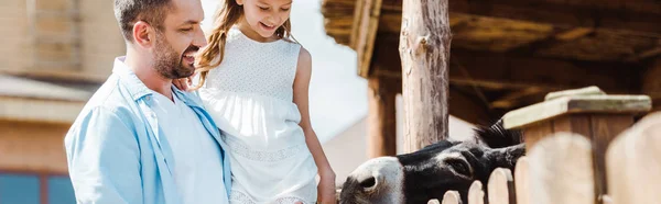 Tiro panorâmico de pai feliz segurando nos braços bonito filha perto de cerca de madeira no zoológico — Fotografia de Stock
