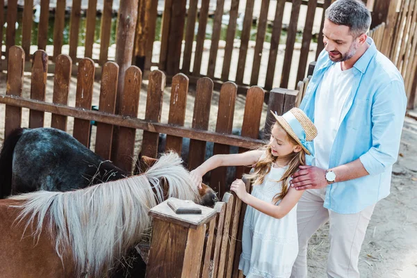 Fröhliches Kind berührt Pony, während es neben Vater im Zoo steht — Stockfoto