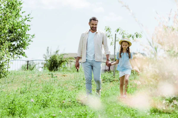 Enfoque selectivo de feliz padre e hija caminando cerca de plantas verdes y tomados de la mano - foto de stock