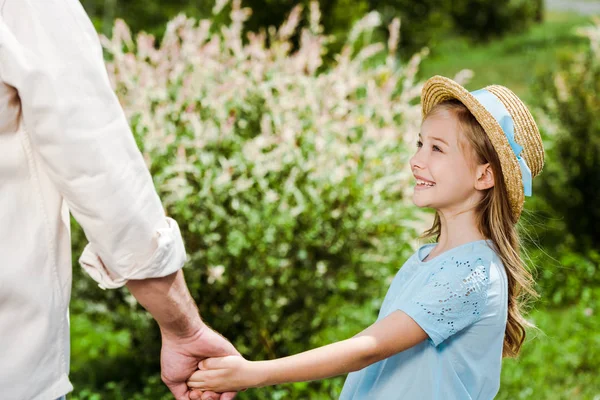 Обрезанный вид отца, держащегося за руки с милой и счастливой дочерью в соломенной шляпе — стоковое фото