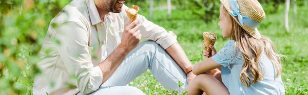 Panoramic shot of father near kid in straw hat with ice cream cone sitting on grass — Stock Photo