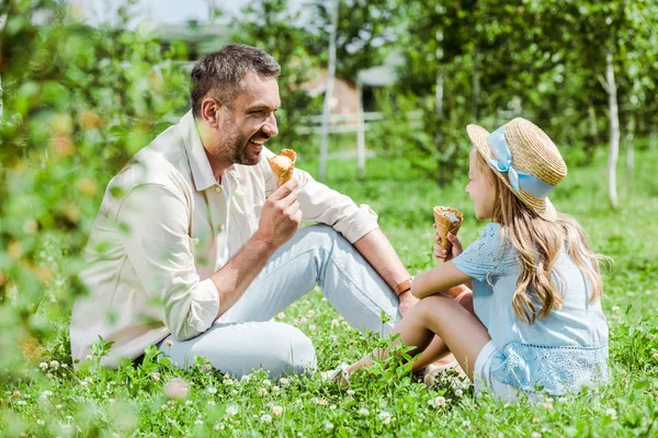 Foco seletivo de pai alegre olhando para criança em chapéu de palha com cone de sorvete sentado na grama — Fotografia de Stock
