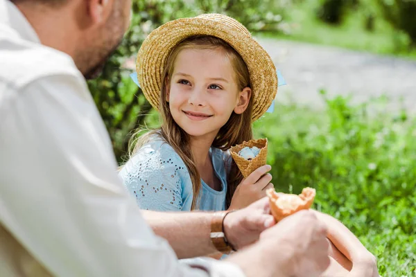 Избирательный фокус веселого ребенка в соломенной шляпе, держащего рожок мороженого и смотрящего на отца — стоковое фото