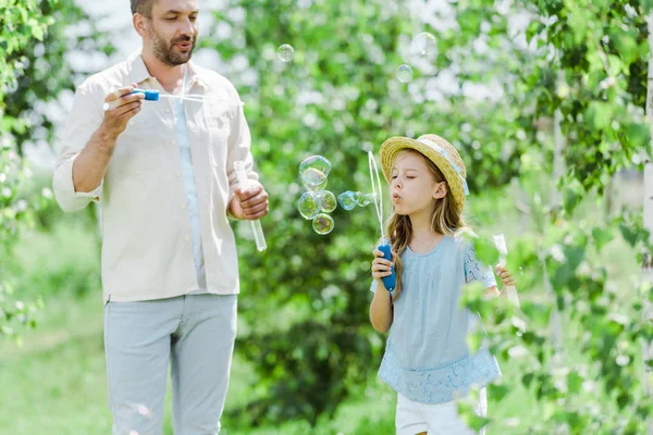 Вибірковий фокус веселого батька, дивлячись на дочку, що дме мильні бульбашки — стокове фото