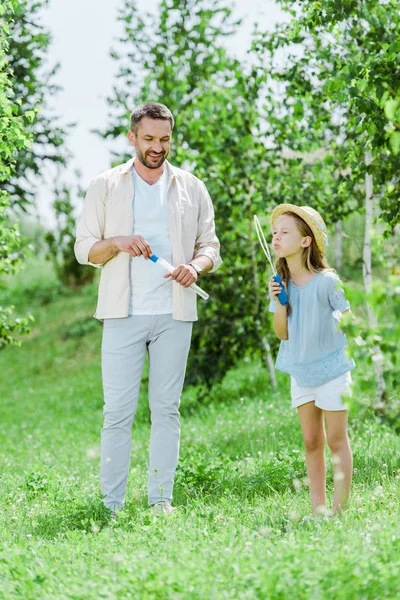 Messa a fuoco selettiva di padre allegro in piedi vicino a figlia che soffia bolle di sapone vicino agli alberi — Foto stock