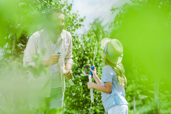 Selective focus of father and daughter in straw hat blowing soap bubbles near trees — Stock Photo
