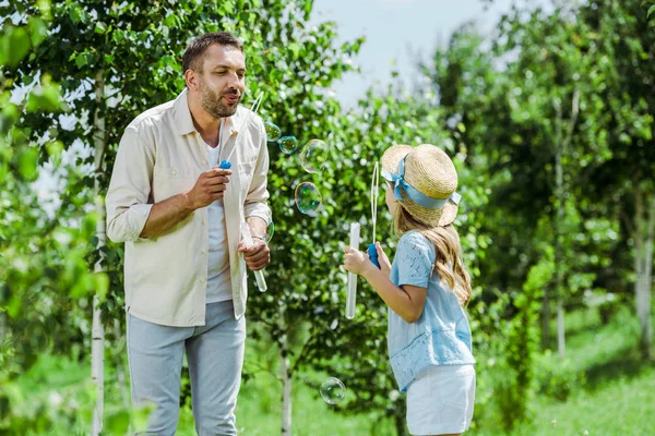 Selective focus of handsome man and daughter in straw hat blowing soap bubbles near trees — Stock Photo