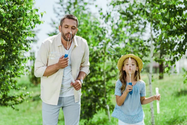 Bello uomo e figlia in cappello di paglia soffiando bolle di sapone vicino agli alberi — Foto stock