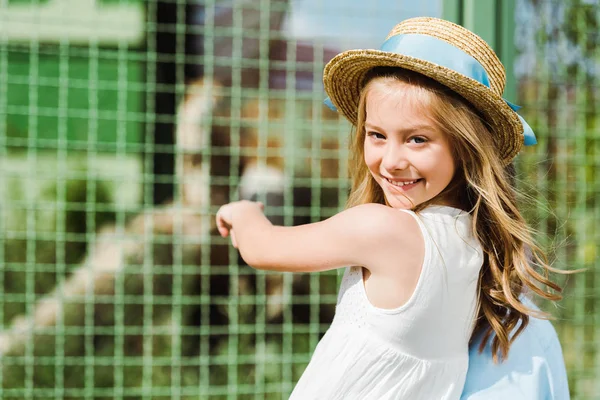 Happy kid in straw hat looking at camera and gesturing near cage in zoo — Stock Photo