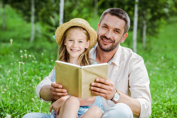 Foco seletivo de pai e filha alegre em chapéu de palha segurando livro e olhando para a câmera — Fotografia de Stock