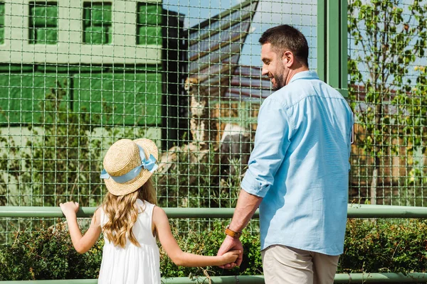 Pai alegre olhando para a filha enquanto segurando as mãos perto da gaiola no zoológico — Fotografia de Stock