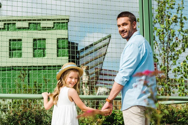 Cheerful father and kid holding hands and looking at camera near cage with leopard in zoo — Stock Photo