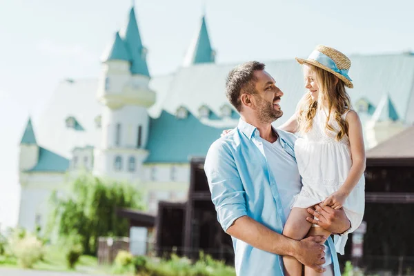 Cheerful father holding in arms cute daughter in straw hat near castle — Stock Photo