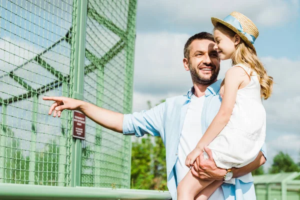 Fröhlicher Mann hält niedliches Kind mit Strohhut in den Armen und zeigt mit dem Finger auf Käfig im Zoo — Stockfoto