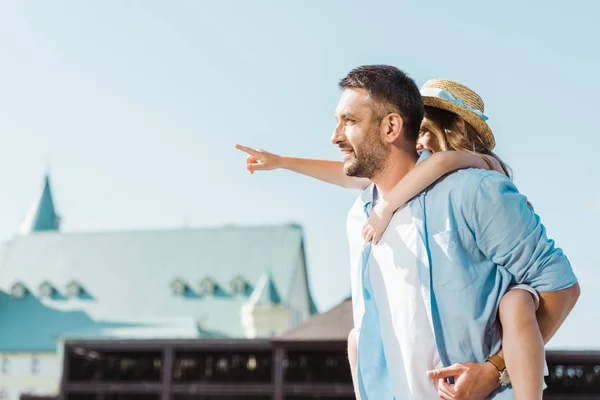 Cheerful man piggybacking daughter pointing with finger near building — Stock Photo