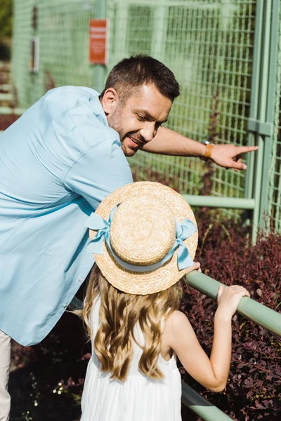 Веселий тато вказує пальцем, дивлячись на дитину в солом'яному капелюсі біля клітки — стокове фото