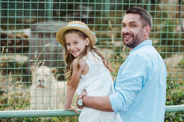 Glücklicher Vater und Tochter blicken in Kamera in der Nähe von Käfig mit wildem Tier im Zoo — Stockfoto