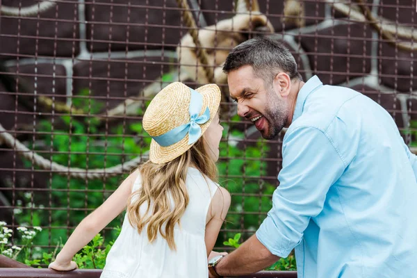 Bambino in cappello di paglia guardando il padre che mostra la lingua vicino alla gabbia con animale selvatico nello zoo — Foto stock
