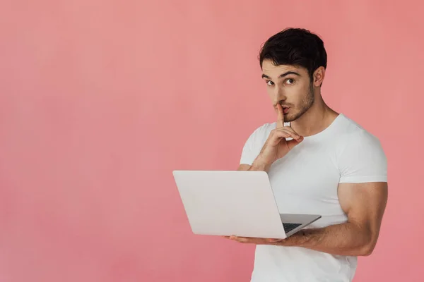 Muscular man in white t-shirt using laptop and showing hush gesture isolated on pink — Stock Photo