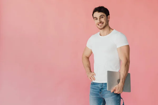 Homem muscular sorridente em t-shirt branca em pé com a mão no quadril e segurando laptop isolado em rosa — Fotografia de Stock