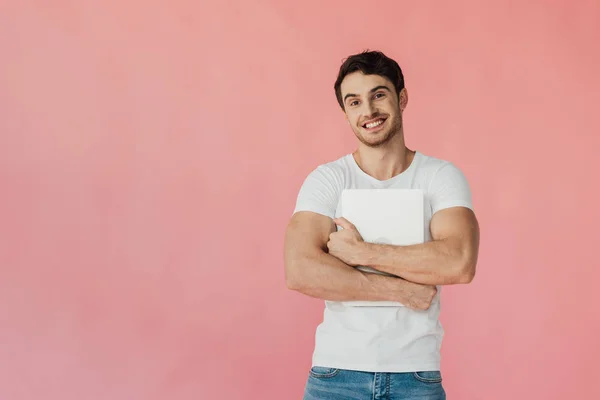 Vista frontal do homem muscular sorridente em branco t-shirt segurando laptop e olhando para a câmera isolada em rosa — Fotografia de Stock