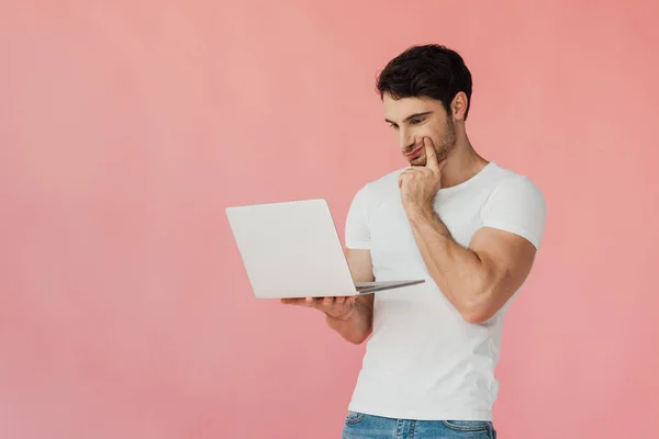 Hombre musculoso pensativo en camiseta blanca usando el ordenador portátil aislado en rosa - foto de stock